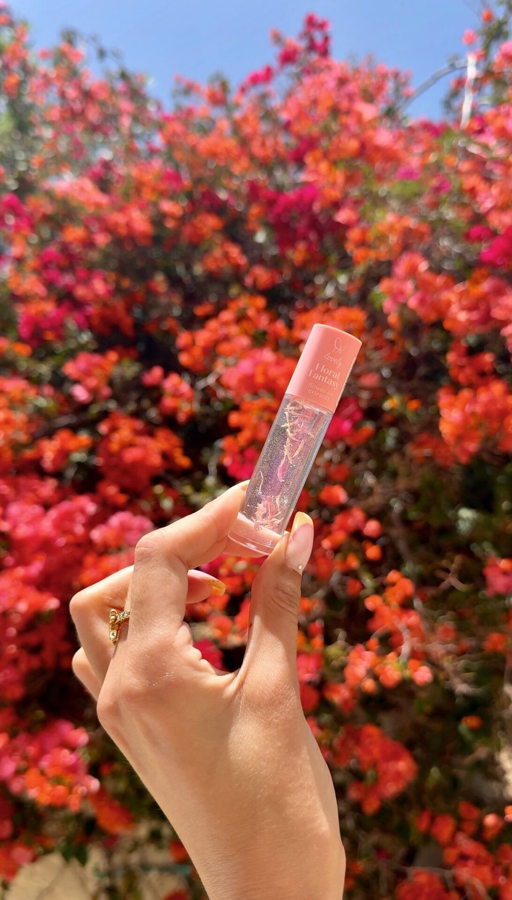 Hand holding a Lovely Floral Fantasy lip oil in front of blooming flowers
